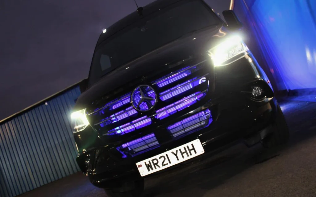 The front of the Mercedes Jet Class Sprinter at night, featuring an illuminated blue grille, bright LED headlights, and the registration plate WR21 YHH.