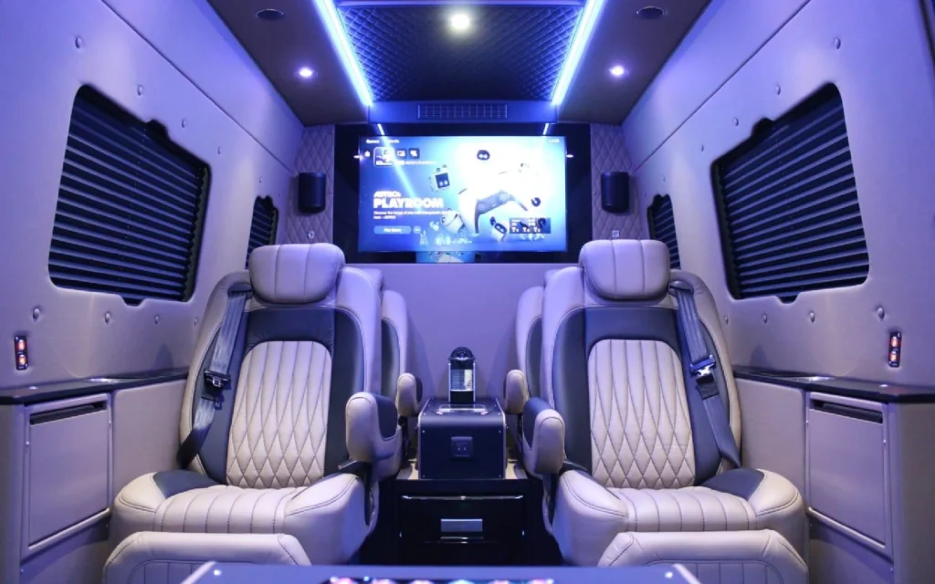 Luxurious interior of the Mercedes Jet Class Sprinter with quilted leather seats, ambient lighting, and a large entertainment screen displaying a PlayStation interface.
